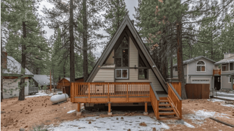 A-frame cabin surrounded by trees, featuring a wooden deck and stairs, with light snow on the ground. Nearby are other houses and a propane tank.