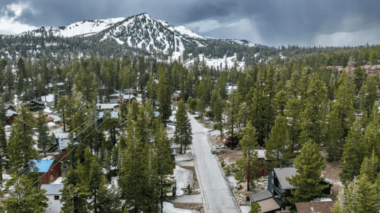 Aerial view of a snowy mountain landscape with pine trees, houses, and a road leading towards the mountains under a cloudy sky.