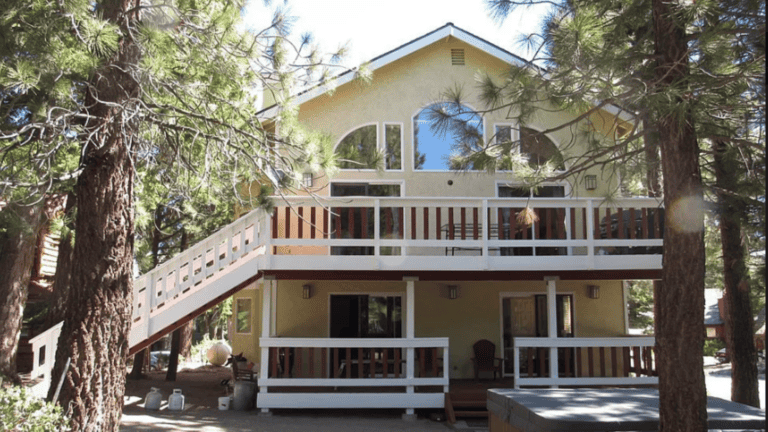 A two-story house with a yellow façade, large windows, and wooden balconies, surrounded by tall pine trees.