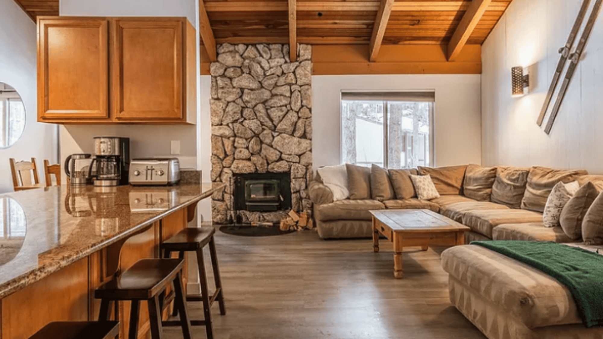 Let's talk Cozy living room with a stone fireplace, wooden beams, and a large sectional sofa. A kitchen counter with stools and appliances is visible on the left. Natural light filters through a window.