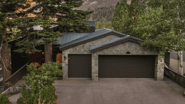 A stone house with a dark roof and two garage doors is surrounded by trees.