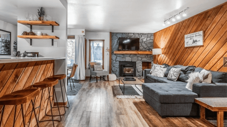 Cozy living room with a stone fireplace, large sectional sofa, wooden panel walls, and a flat-screen TV. Kitchen area with bar stools and open shelving visible to the left.