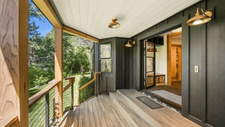 Covered porch with wooden floorboards, overlooking a green yard. Black exterior walls, glass double doors, and modern wall lights.