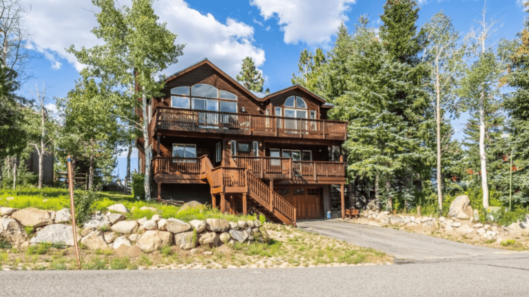 A large wooden cabin with multiple balconies is surrounded by trees and set on a stone foundation. A paved driveway leads to the entrance.