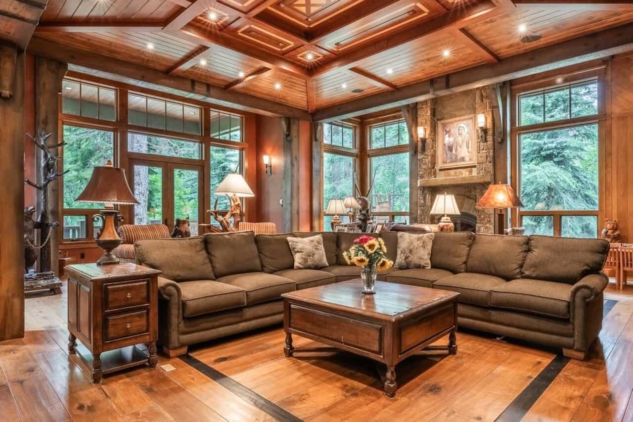 Let's talk Spacious living room with brown sectional sofa, wooden coffee table, large windows, stone fireplace, wood-paneled ceiling, and forest view outside.