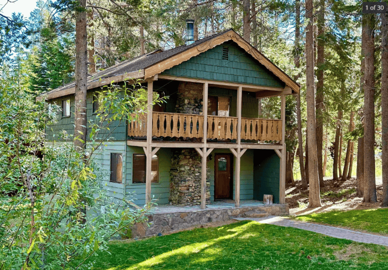 Two-story green wooden cabin with a stone chimney, balcony, and covered porch, surrounded by tall trees and grass in a forested area.