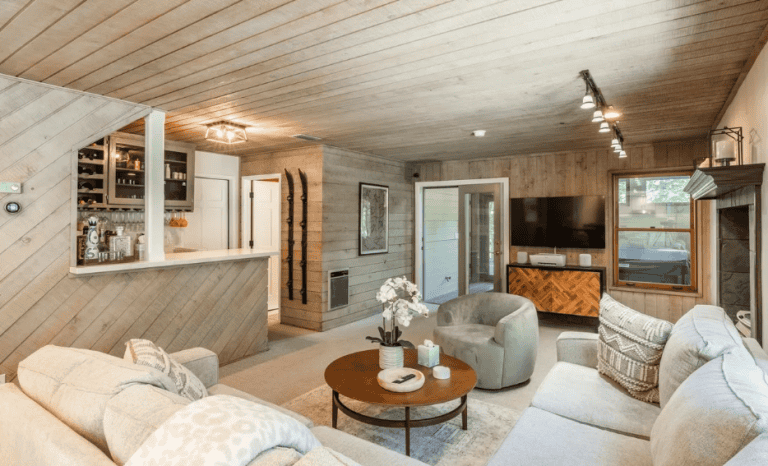 A cozy living room with wood-paneled walls and ceiling, a round coffee table, upholstered seating, a wall-mounted TV, a fireplace, and a small bar area with shelves and wine storage.