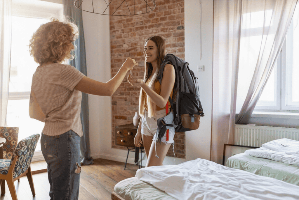 A woman hands a key to a young woman with a backpack inside a bright, tidy room with two beds and large windows.