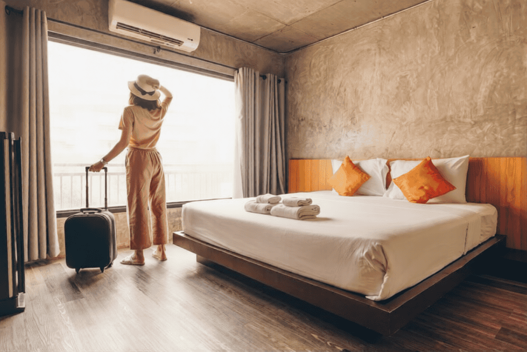 A person with a suitcase stands by a large window in a modern hotel room with a double bed, white linens, and two orange pillows.