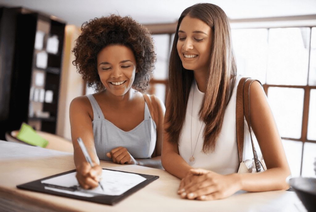 Two women stand at a counter; one is smiling while signing a document on a clipboard, and the other looks on, also smiling.