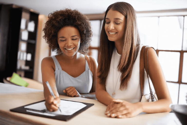 Two women stand at a counter; one is smiling while signing a document on a clipboard, and the other looks on, also smiling.