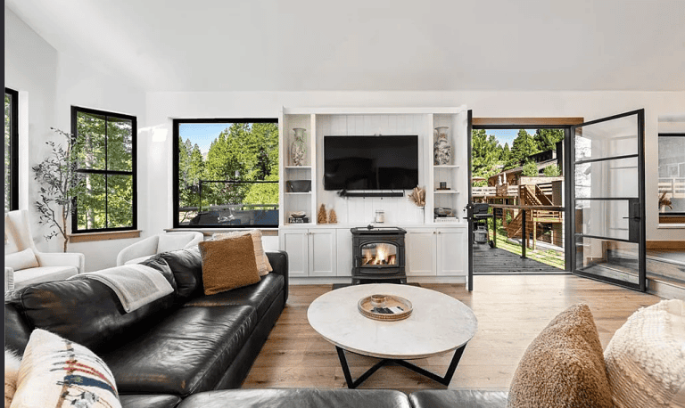 Modern living room with black leather sofa, round coffee table, fireplace, built-in shelves, wall-mounted TV, large windows, and open doors leading to an outdoor deck with forest views.