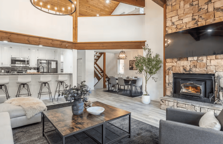 Spacious open-concept living area with stone fireplace, modern kitchen with bar stools, dining table, vaulted ceiling, and large indoor plant.