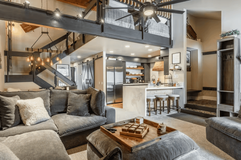 Modern open-plan living area with grey sectional sofa, wooden coffee table, kitchen with bar stools, lofted beds, and exposed beams. Neutral tones and natural light fill the space.