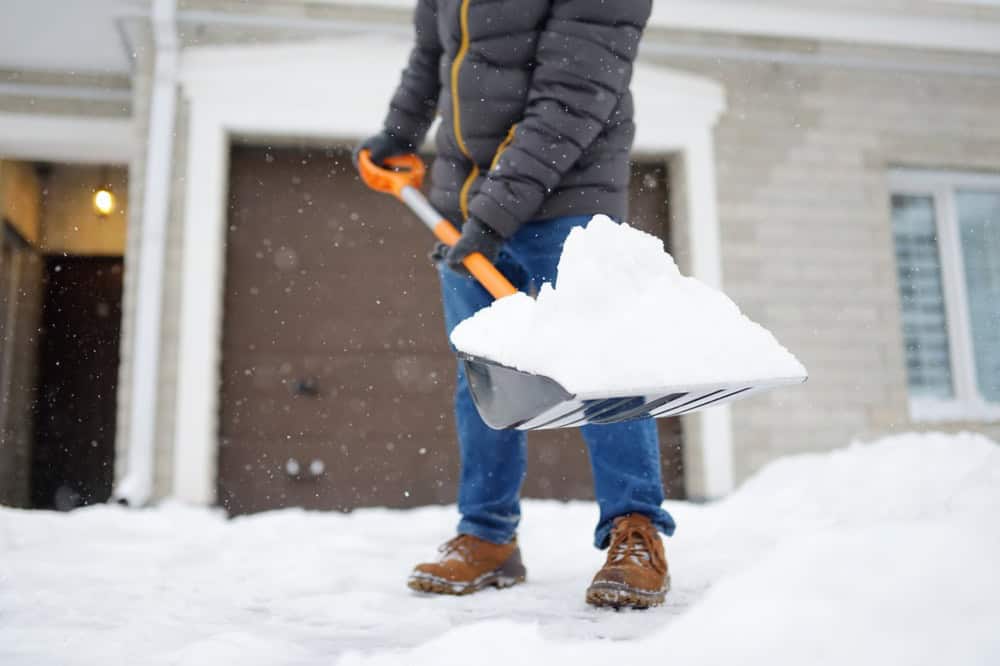 A person in a puffer jacket and jeans shoveling snow in front of a house.
